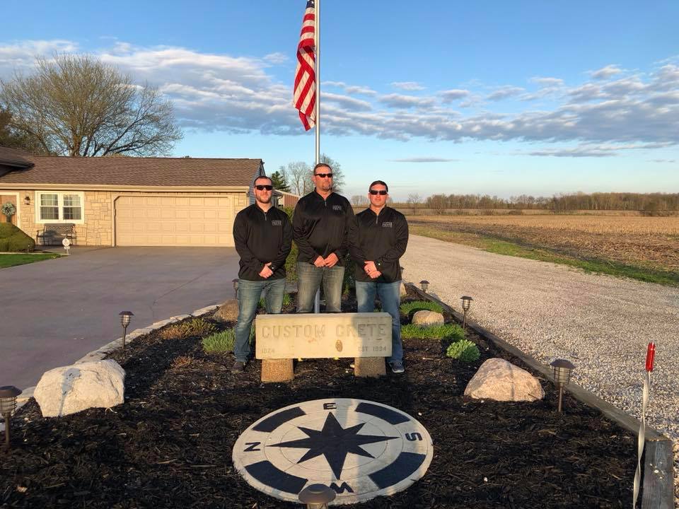The Custom Crete crew standing behind their engraved monument sign and stamped compass-rose medallion with an American flag on a rural Central Ohio property