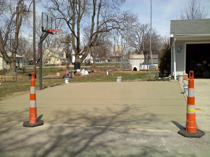 Freshly poured tan concrete pad for a backyard basketball court, marked off with traffic cones beside a basketball hoop.