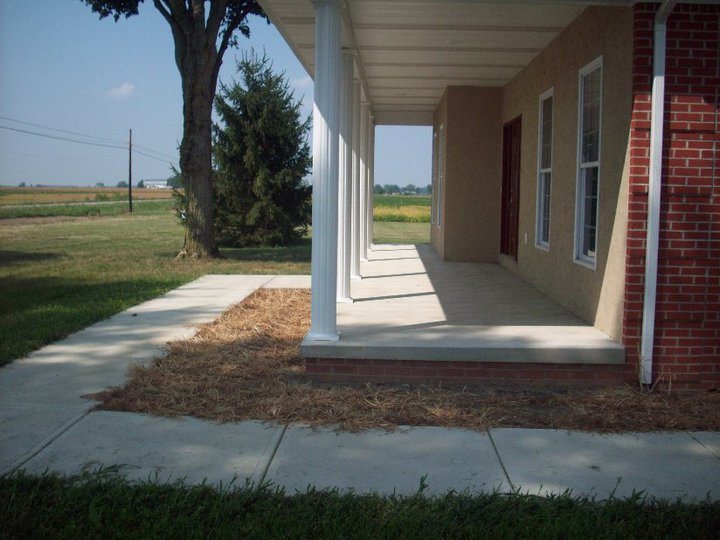 A finished plain concrete covered porch and connecting sidewalk on a brick and stucco rural home.
