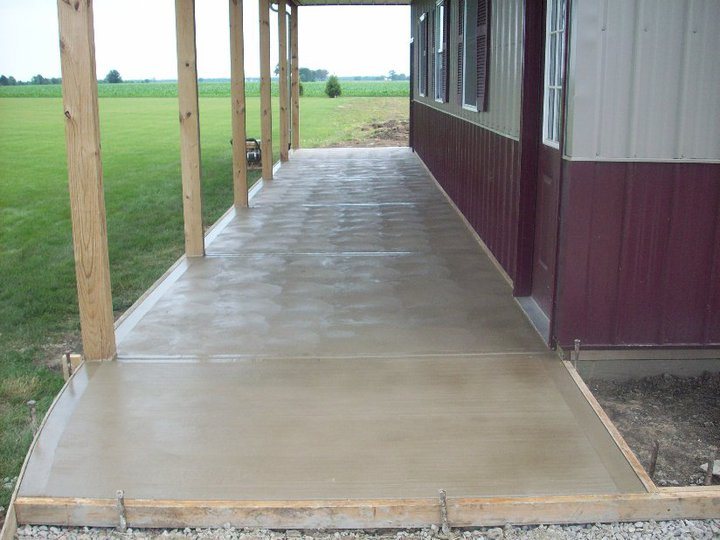 A finished plain concrete porch slab running along the side of a maroon pole barn under a post-supported roof.