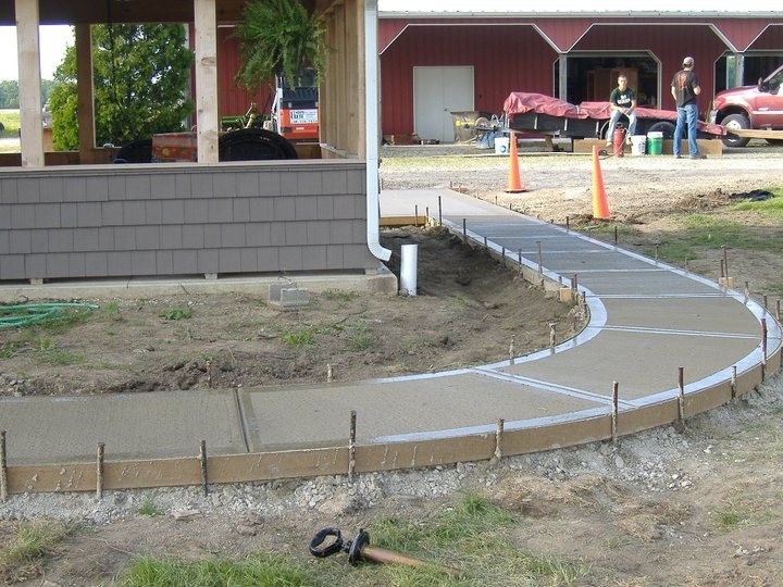 Freshly poured curved concrete walkway still in its forms in front of a farm building with a red barn behind.