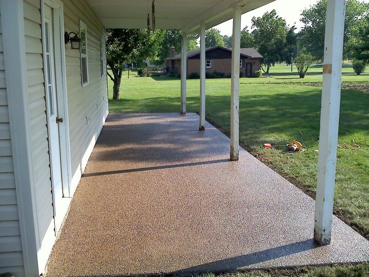 Tan exposed aggregate concrete floor on a covered front porch of a white sided house.