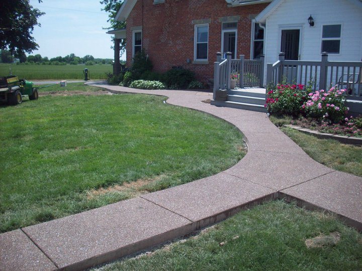 Curved exposed aggregate concrete walkway with stained borders leading to a brick farmhouse porch lined with rose bushes.