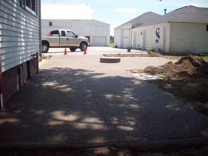 A finished exposed-aggregate concrete driveway between rural houses with a pickup truck and safety cones.