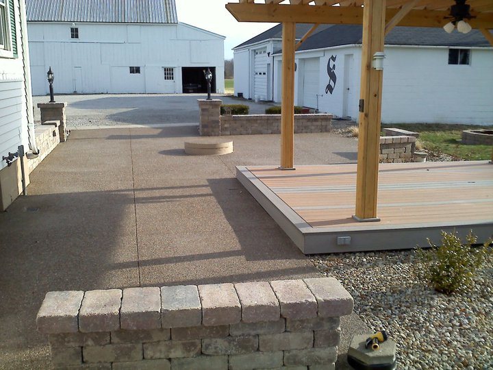 Exposed aggregate concrete patio with stone seat walls and a wood pergola beside a white barn on a rural property.