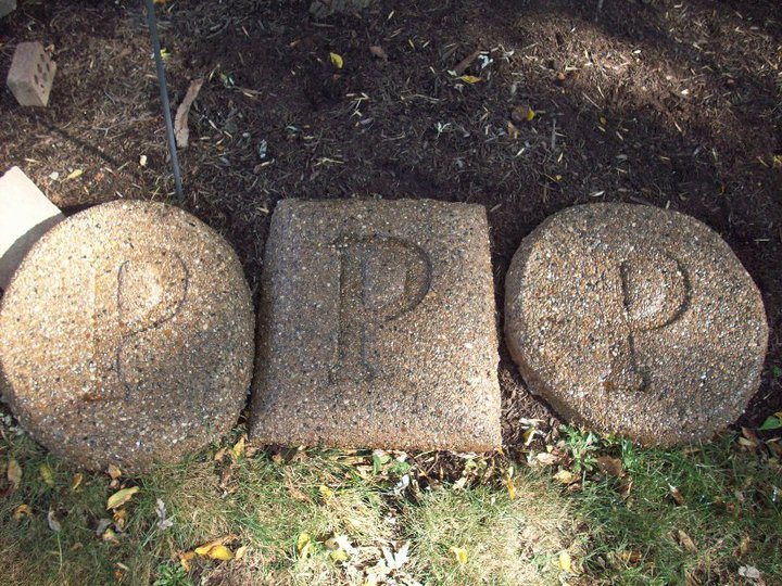 Three exposed-aggregate concrete stones engraved with the letter P set in a mulched landscape bed.