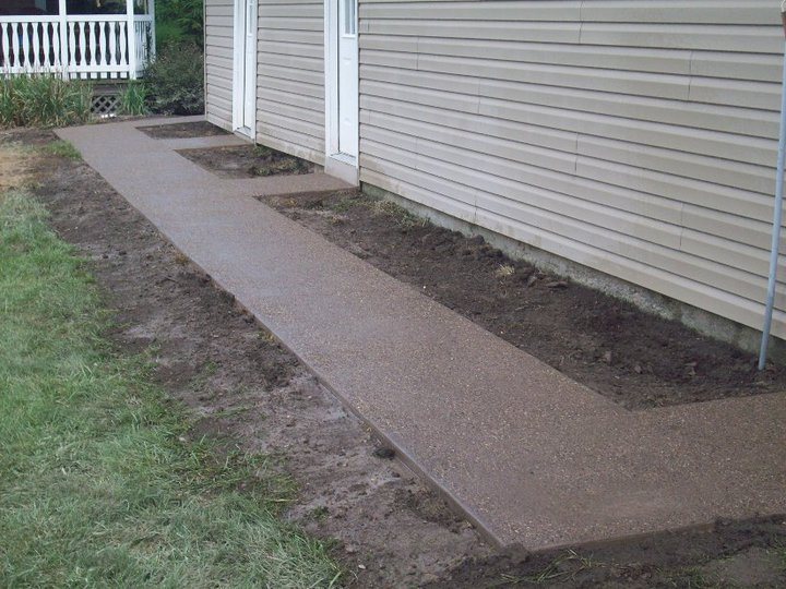 A finished tan exposed-aggregate concrete sidewalk with step landings runs along the side of a tan-sided house.