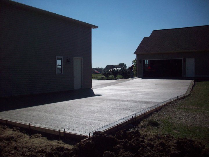 Freshly poured broom-finished concrete parking pad between a gray outbuilding and a garage, with wood forms still in place.