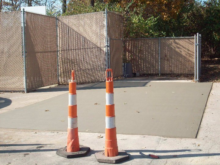 A freshly poured rectangular concrete pad inside a chain-link fenced enclosure marked off with traffic cones.