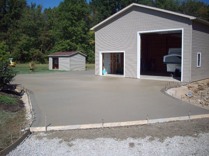 Freshly poured smooth gray concrete apron slab with edge forms in front of a tan pole-barn garage.