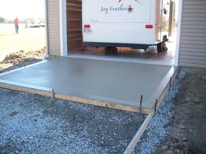 Freshly poured smooth concrete pad with wood forms at the doorway of a sided outbuilding housing a Jay Feather camper, bordered by gravel.