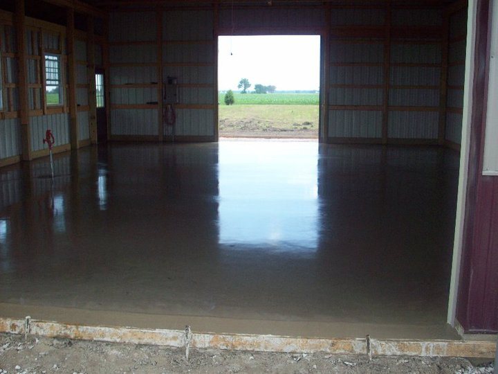 A smoothly troweled, freshly finished concrete floor poured inside a metal barn open to a cornfield.