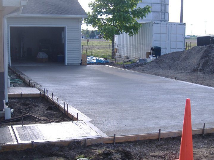 Newly poured concrete garage apron slab still in forms beside a grain bin and shipping container in a rural setting.