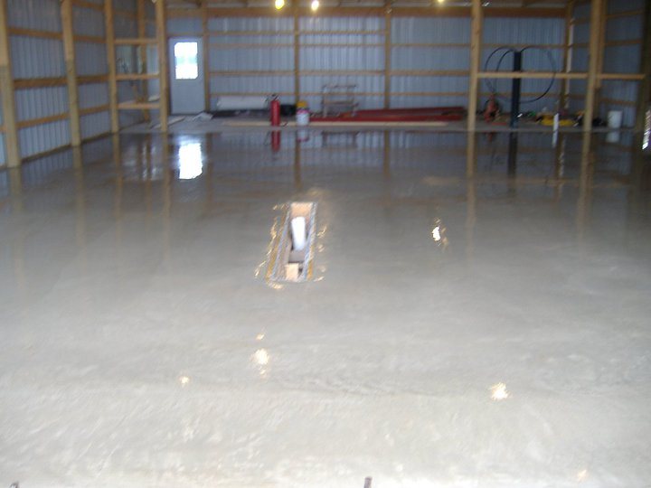 Freshly poured and troweled wet concrete floor inside a metal pole building with a floor drain opening.