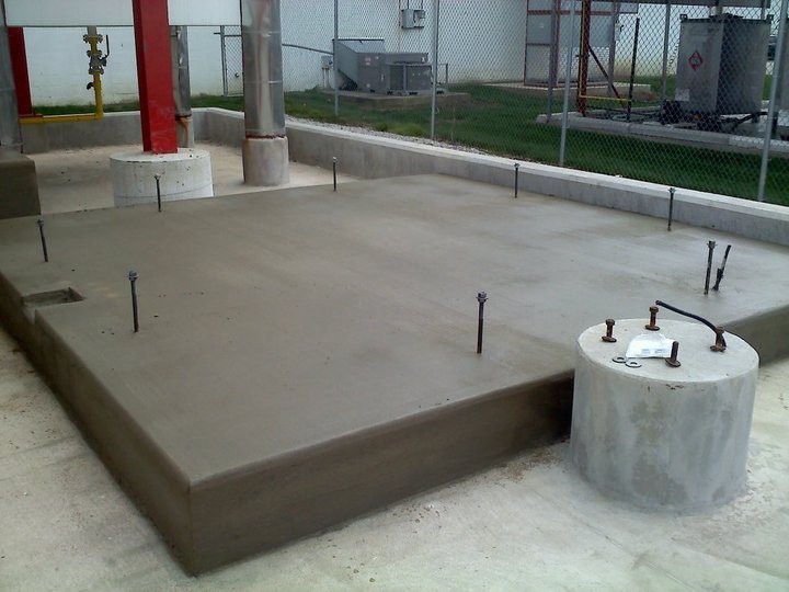Freshly poured concrete equipment pad with protruding anchor bolts at a fenced industrial facility.