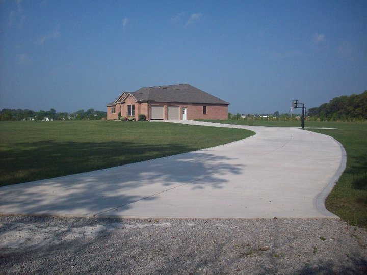 A long curving plain concrete driveway leading to a brick ranch home across an open rural lawn.