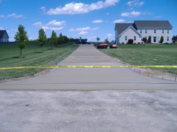 Long freshly finished plain concrete driveway running uphill to a two-story home, marked off with caution tape.