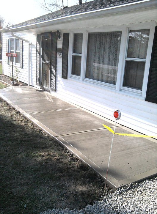 Freshly poured plain broom-finished concrete walkway running along the front of a white sided house.
