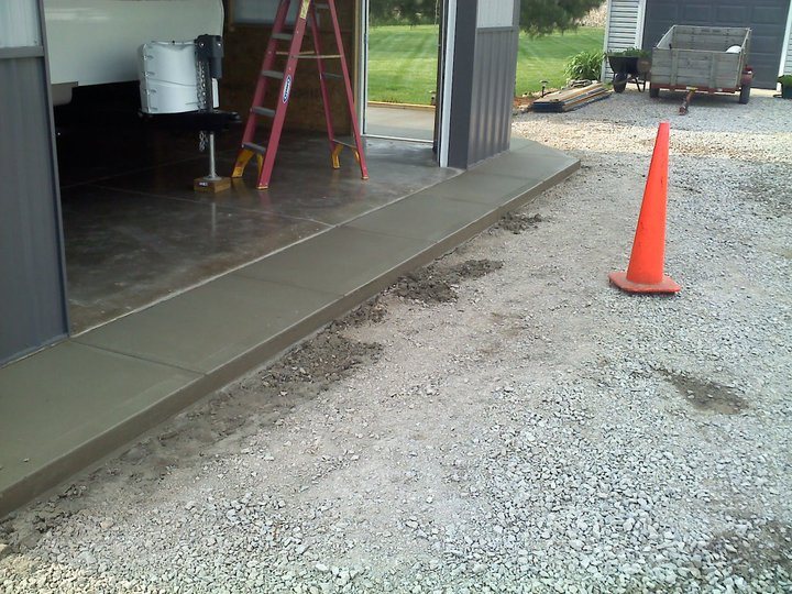 Plain broom-finished concrete apron poured at the entrance of a metal pole barn next to gravel.