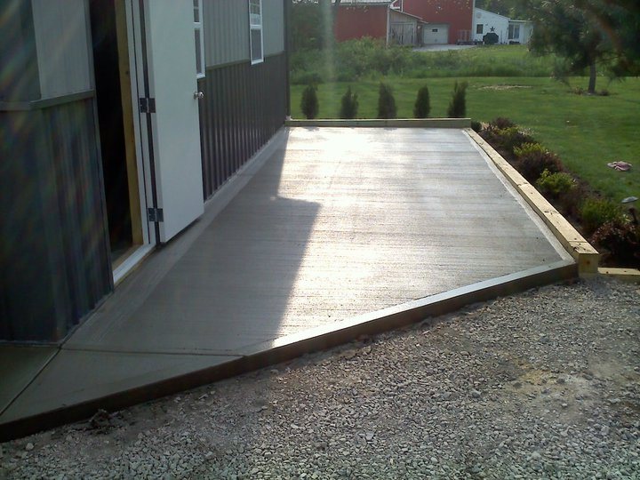 Newly poured plain concrete pad with wood forms at the doorway of a metal storage shed.
