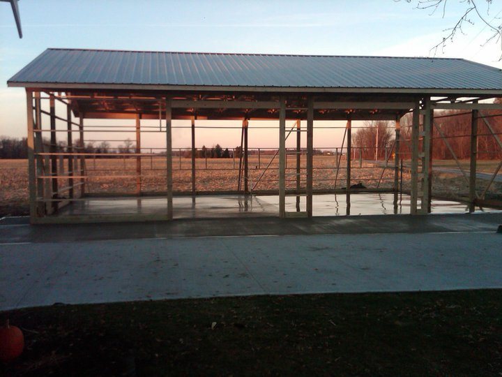 A completed plain concrete slab poured under a finished metal-roofed pole barn at dusk in a rural field.