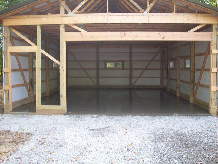 Newly poured smooth concrete floor slab inside a wood-framed pole-barn garage under construction surrounded by gravel.