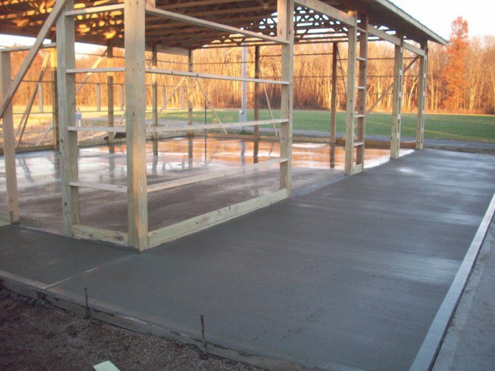 A newly poured and finished concrete slab with formed edges inside an open wood-framed pole barn in a rural setting.