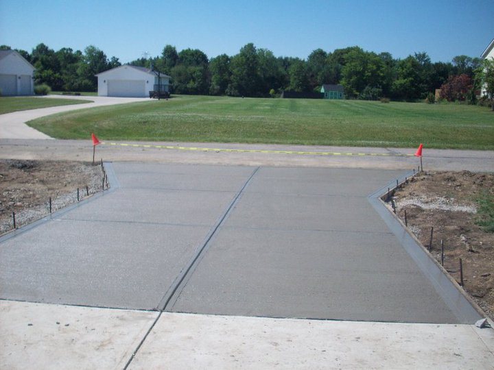 A finished plain concrete driveway apron at the road edge, freshly cured with marker flags and caution tape.