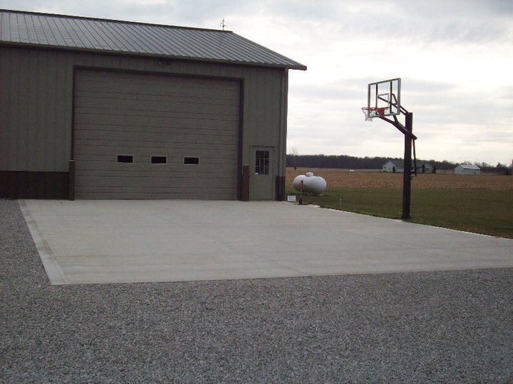 A large plain concrete apron pad in front of a tall metal pole building with a basketball hoop alongside in a rural setting.
