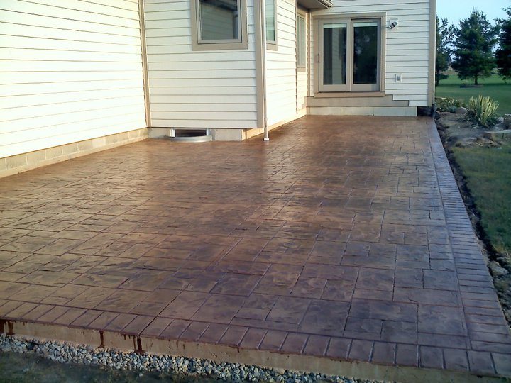 Sealed brown ashlar-pattern stamped concrete patio with a darker border along the back of a sided house.