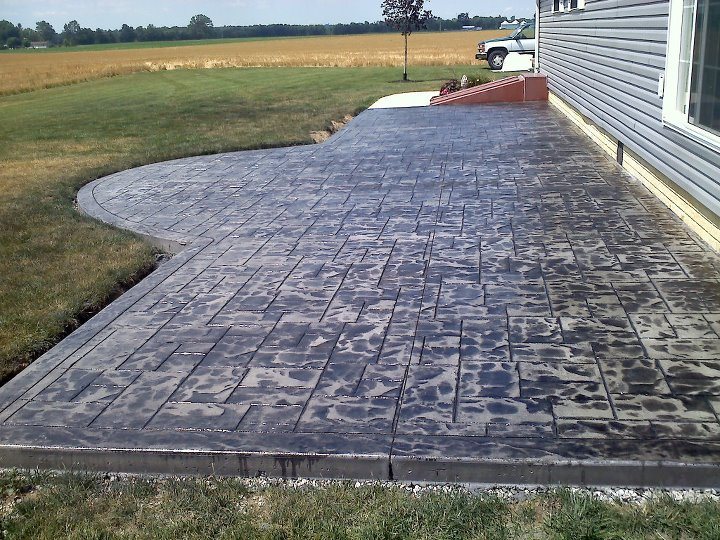 A finished dark gray ashlar-pattern stamped concrete patio along a home's siding overlooking a rural field.
