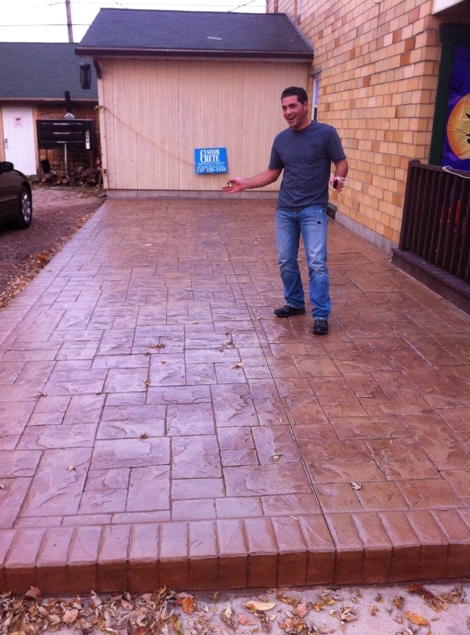 Man stands beside a tan stamped concrete patio with an ashlar slate pattern and brick-stamped border, holding a Custom Crete yard sign.