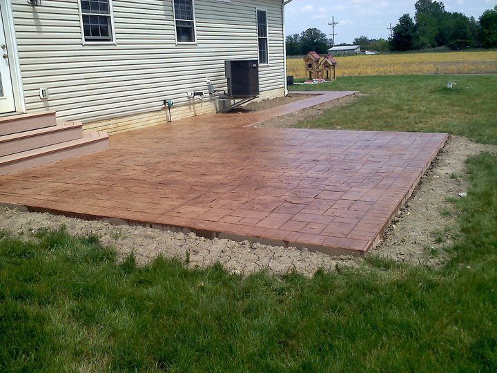 Terracotta-stained ashlar-pattern stamped concrete patio and connecting walkway behind a tan sided house.