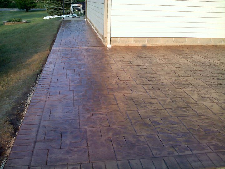 Reddish-brown ashlar-pattern stamped concrete patio and connecting walkway wrapping around a sided house.