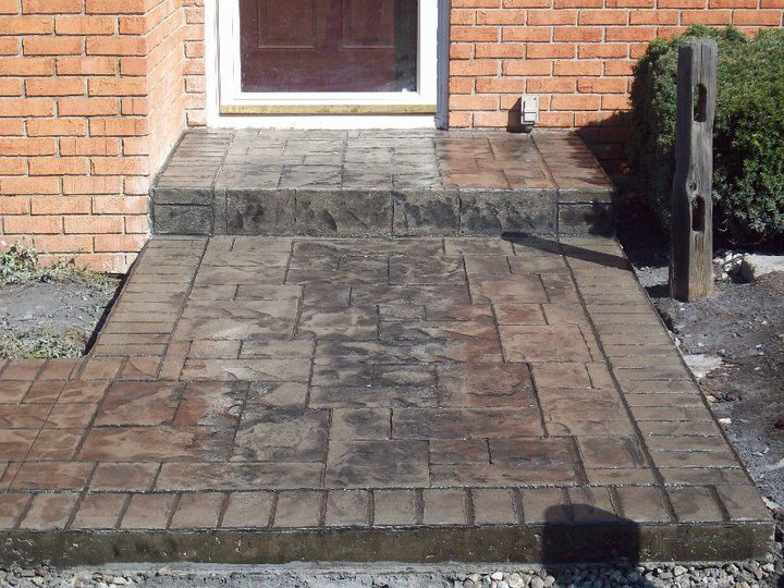 Tan stamped concrete porch and step with an ashlar pattern and brick-stamped border against a brick house entry.