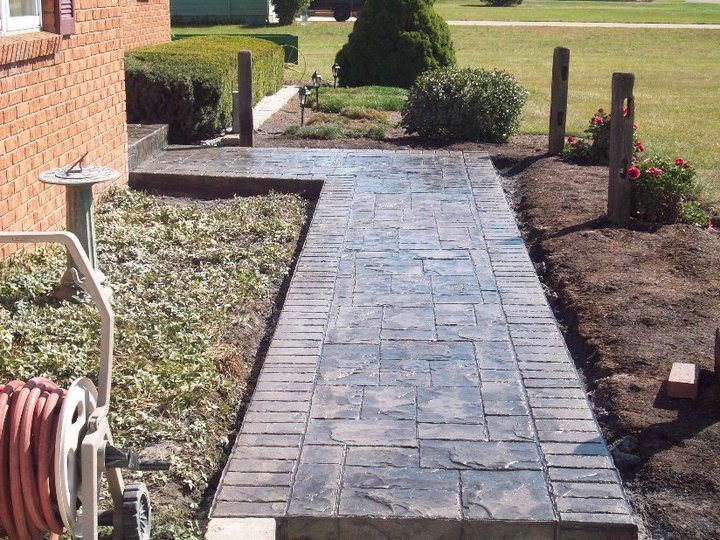 Slate-gray stamped concrete walkway with an ashlar pattern and brick-stamped border leading to a brick house with flower beds.