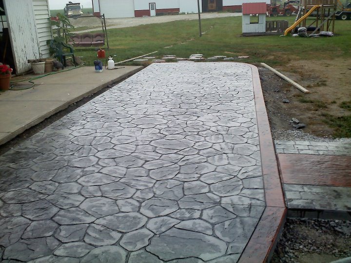 A gray random-stone stamped concrete patio with red-stained border set in a rural farmyard.