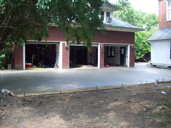A freshly poured concrete driveway apron curing in front of an open three-car brick garage with wood forms along the edge.