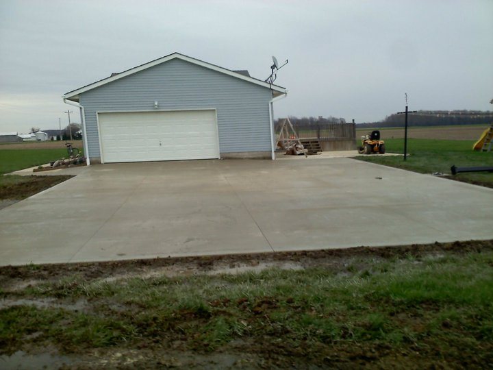 Wide plain gray concrete driveway in front of a detached blue garage on a rural lot.
