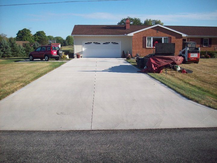 A wide finished plain concrete driveway in front of a two-car garage on a brick ranch home with a work trailer parked beside.