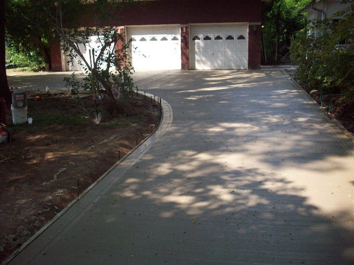 A finished plain concrete driveway leading to a three-bay garage on a brick home, shaded by trees.
